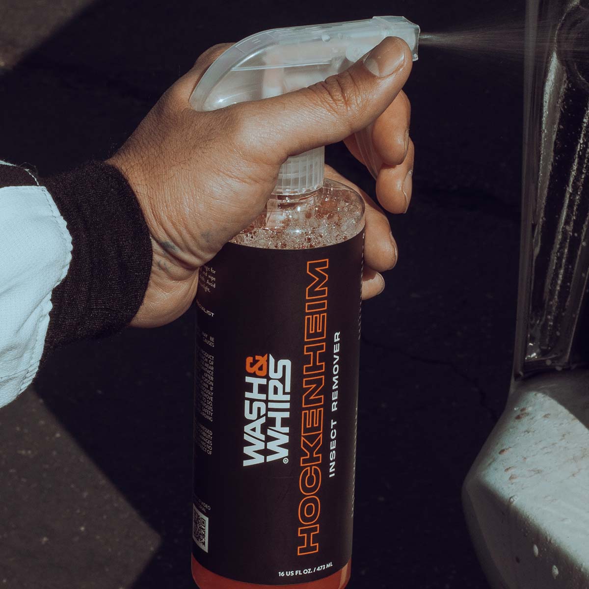 Hand holding a bottle of Hockenheim Wash & Whips insect remover, spraying it onto a surface.