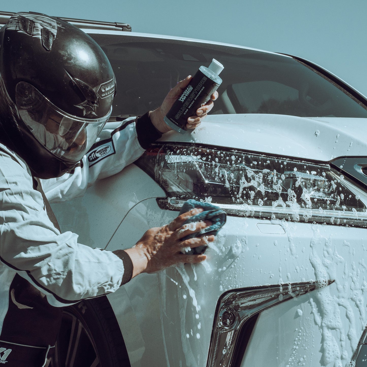 Person cleaning a car with a sponge and bottle, wearing a helmet and white jacket.