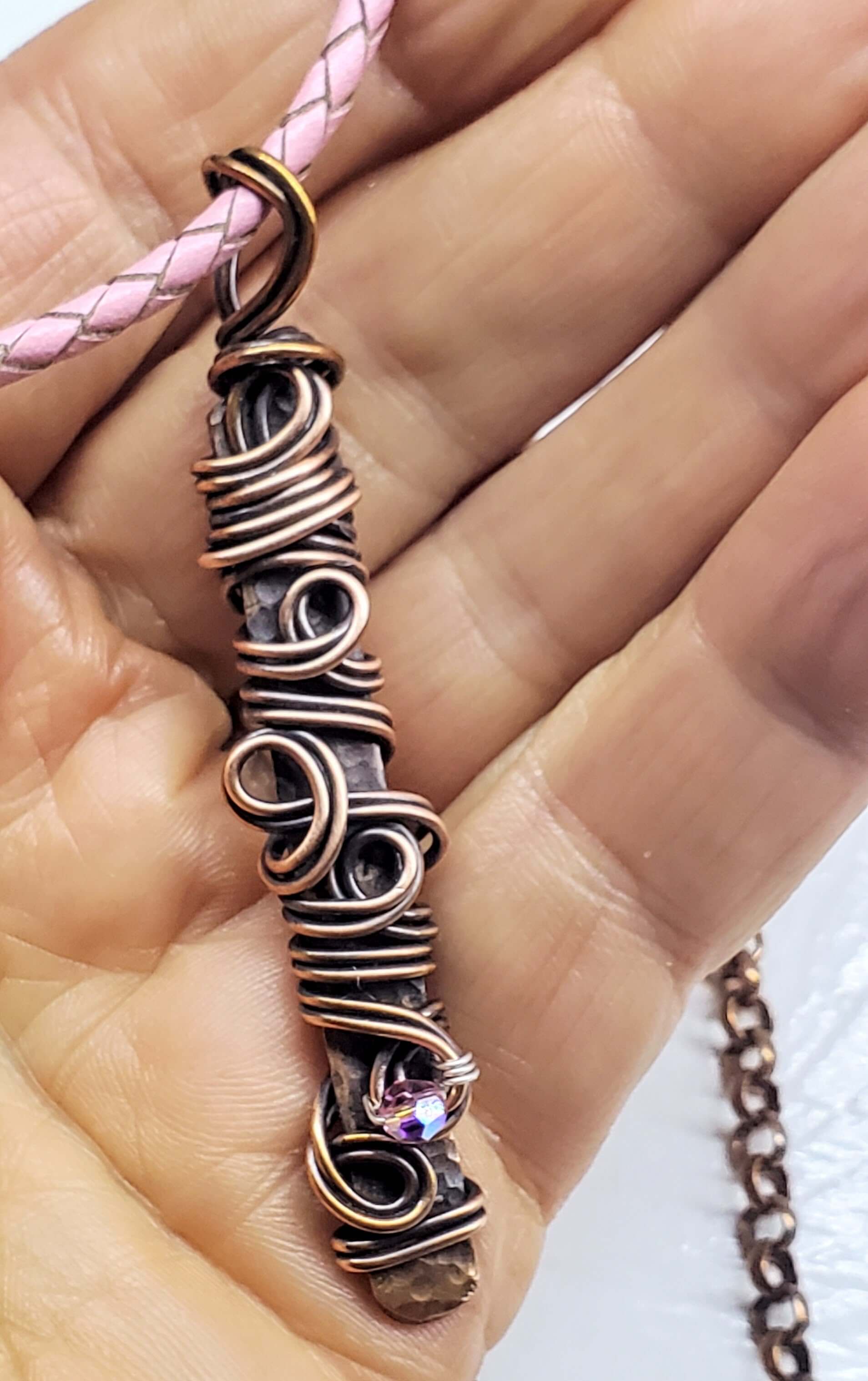Hand holding a intricately designed copper necklace with pink braided cord.