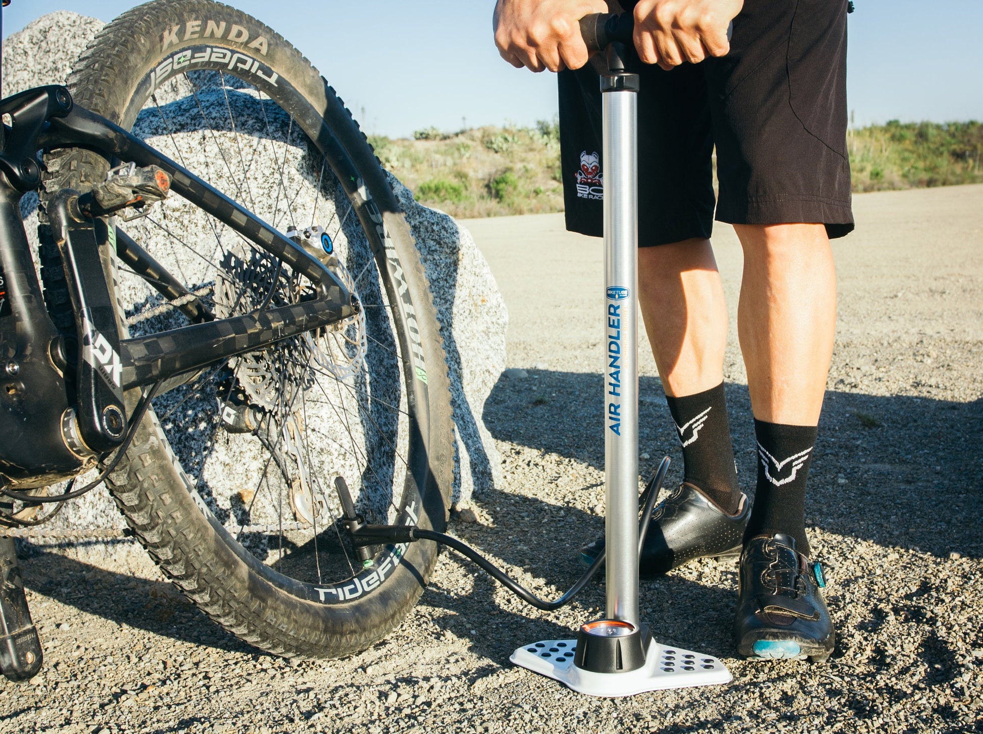 Person inflating a bicycle tire with a pump on a gravel road.