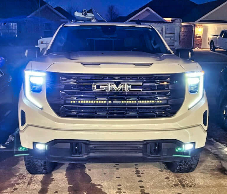White GMC truck with illuminated headlights in a residential driveway.