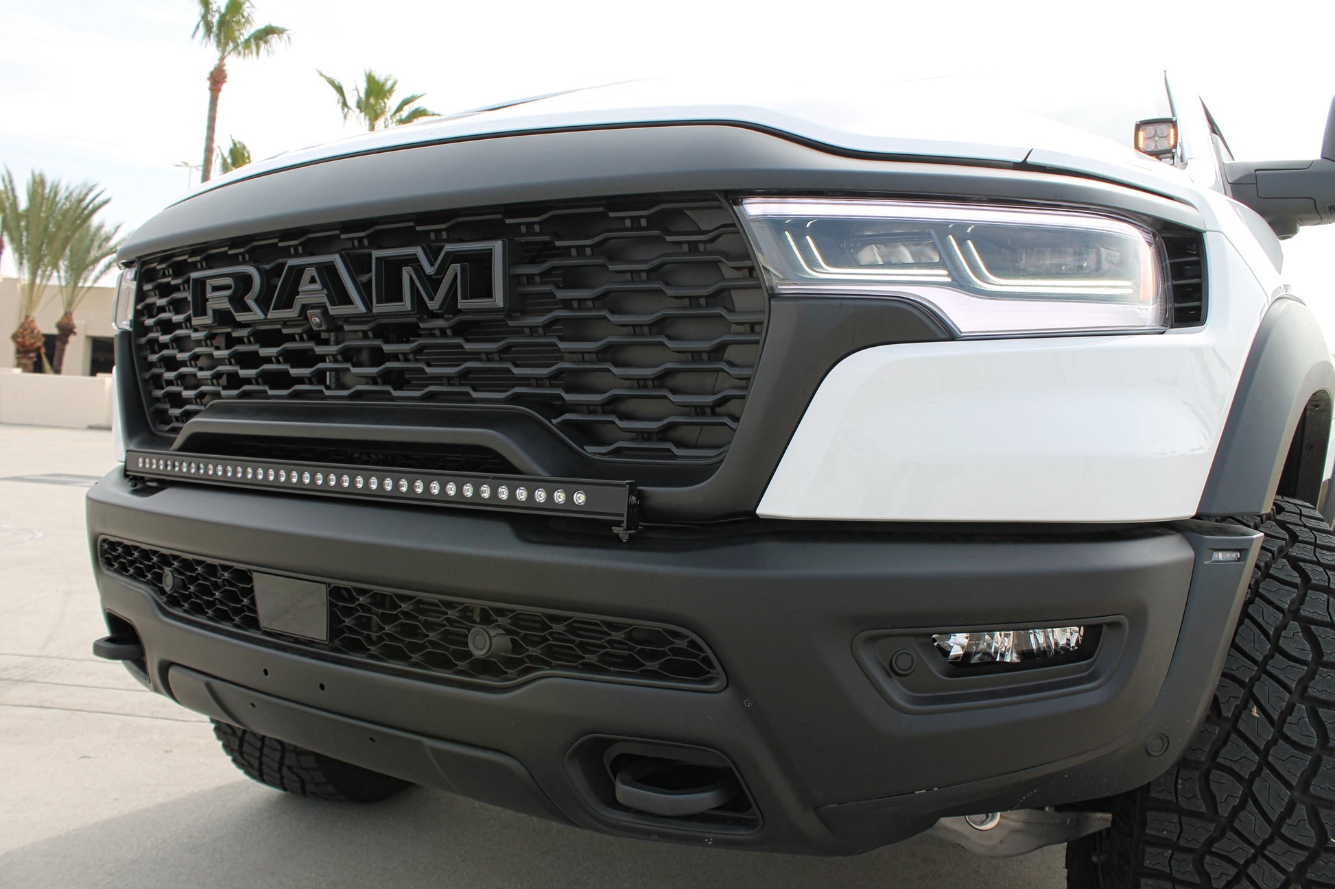 Close-up of a white Ram truck's front grille with light bar and palm trees in the background.