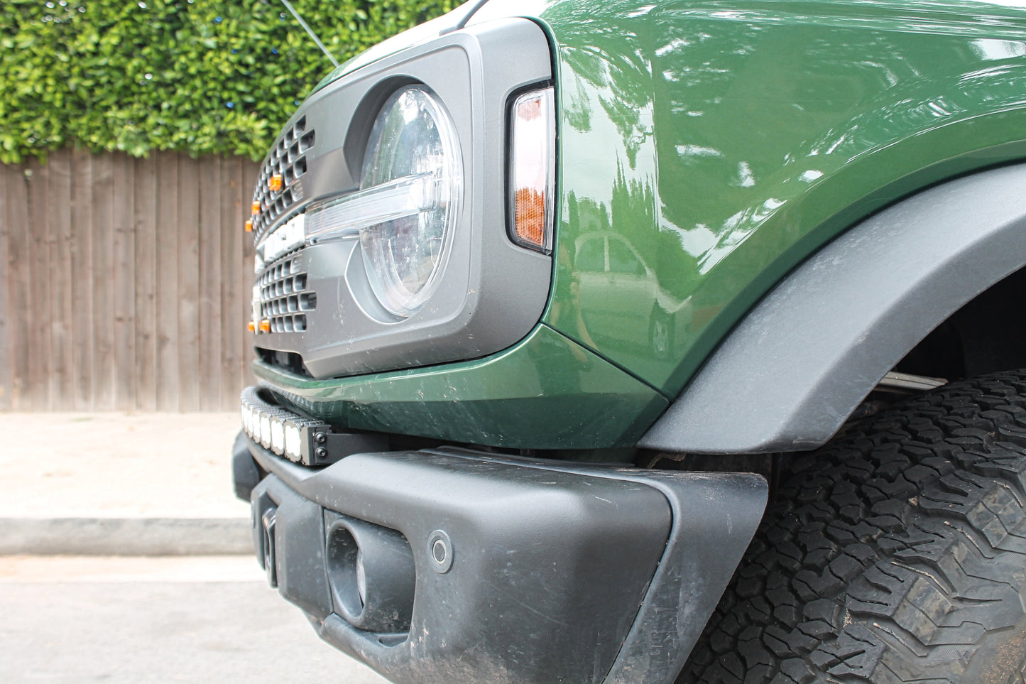 Close-up of a green Bronco truck's front bumper, headlight and LED Light Bar with a wooden fence and greenery in the background.