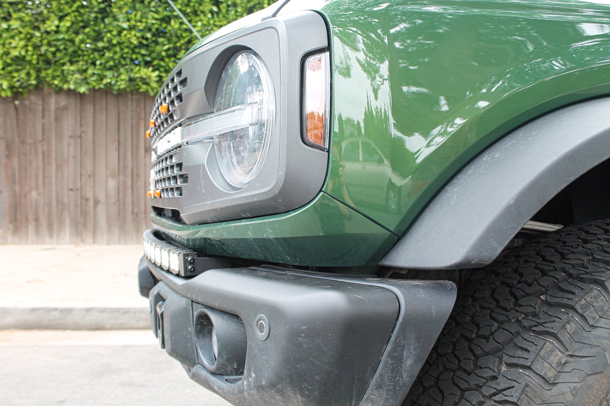 Close-up of a green Bronco truck's front bumper, headlight and LED Light Bar with a wooden fence and greenery in the background.