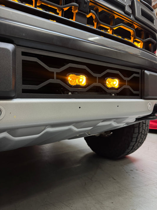Close-up of a Ford Raptor Gen 3 Refresh Guard Plate on a front bumper with amber lights on a concrete floor.