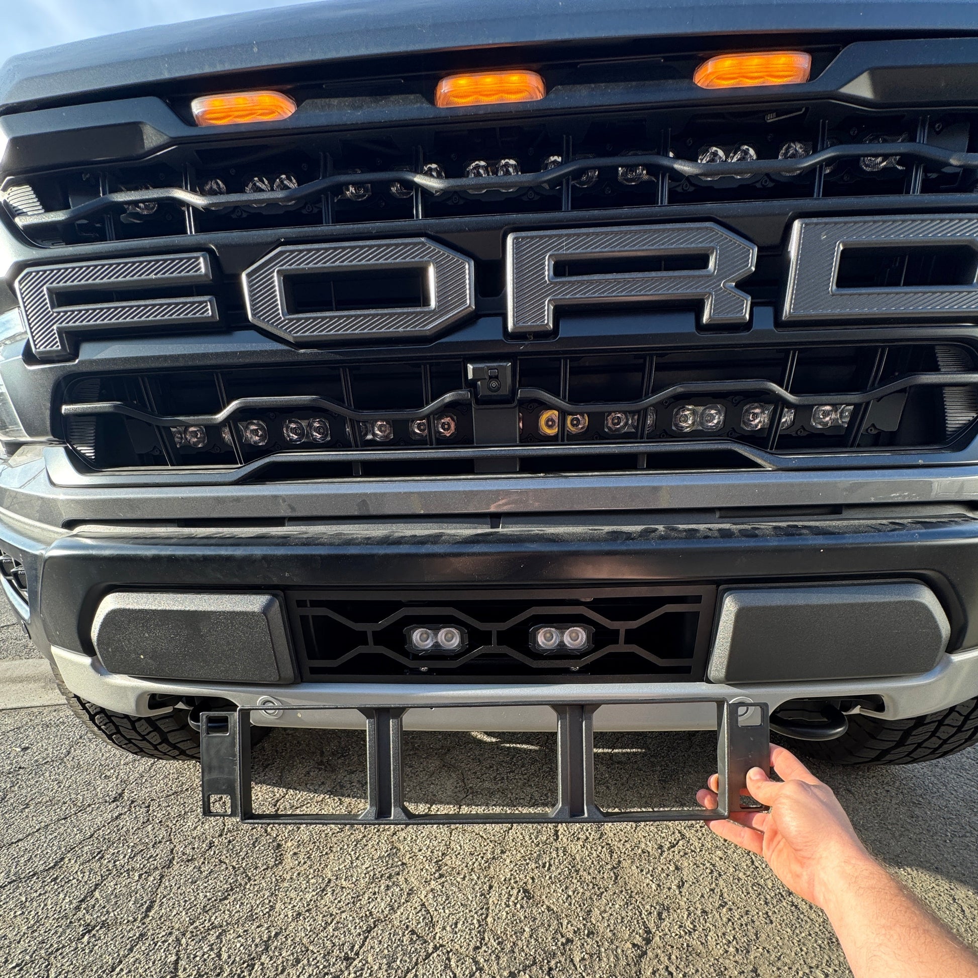 Person holding a black grille guard in front of a Ford truck. Ford Raptor Gen 3 Refresh Guard Plate
