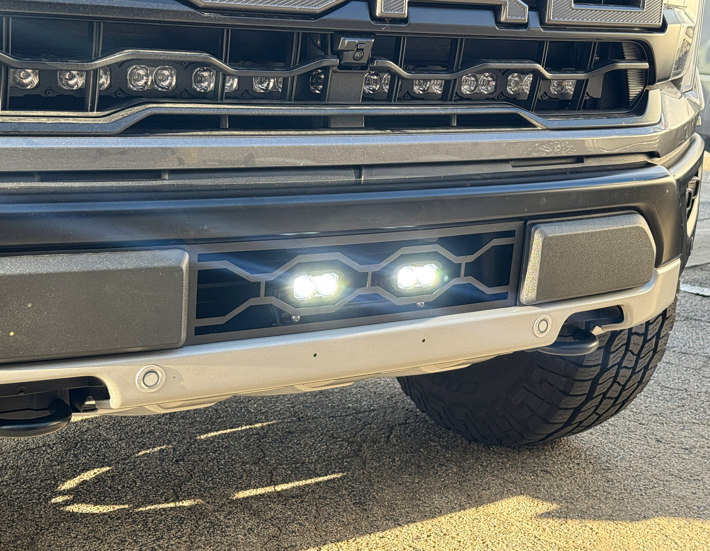 Close-up of a vehicle's front bumper with Ford Raptor Gen 3 Refresh Guard Plate with fog lights on a road.