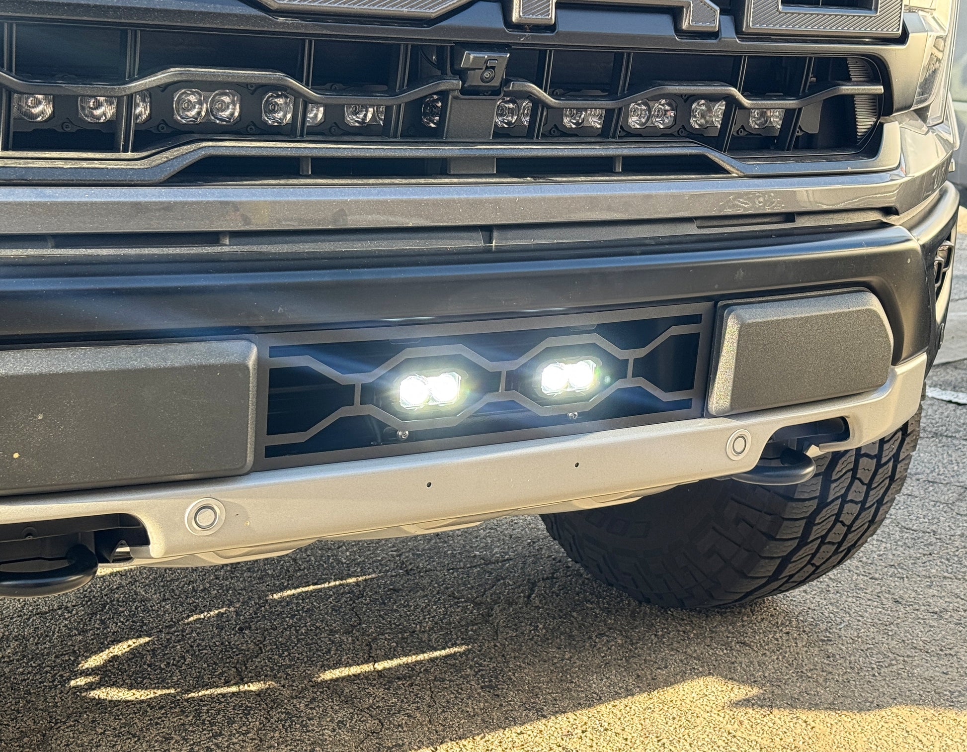 Close-up of a vehicle's front bumper with Ford Raptor Gen 3 Refresh Guard Plate with fog lights on a road.