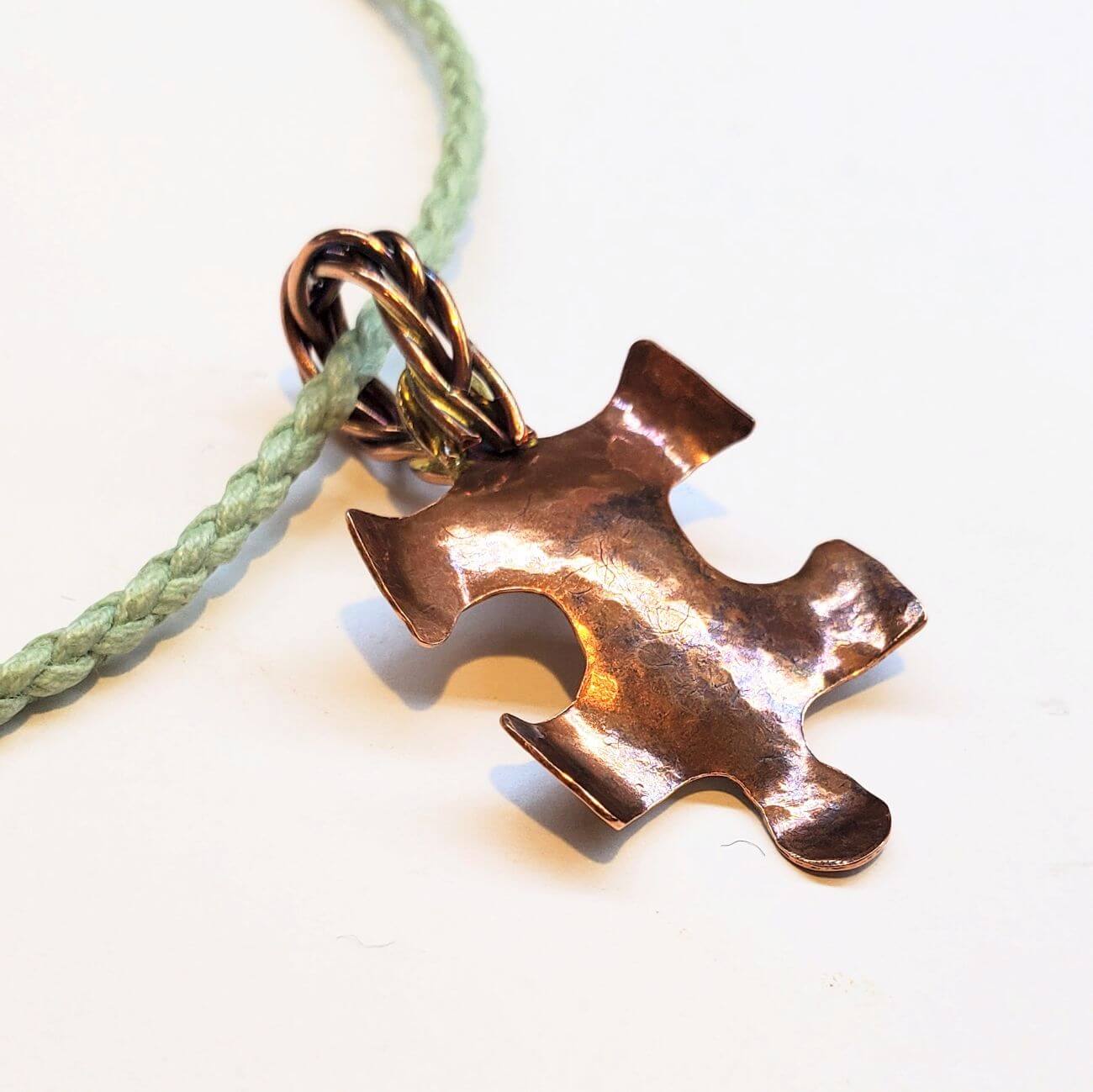 Bronze puzzle piece pendant on a green braided cord against a white background