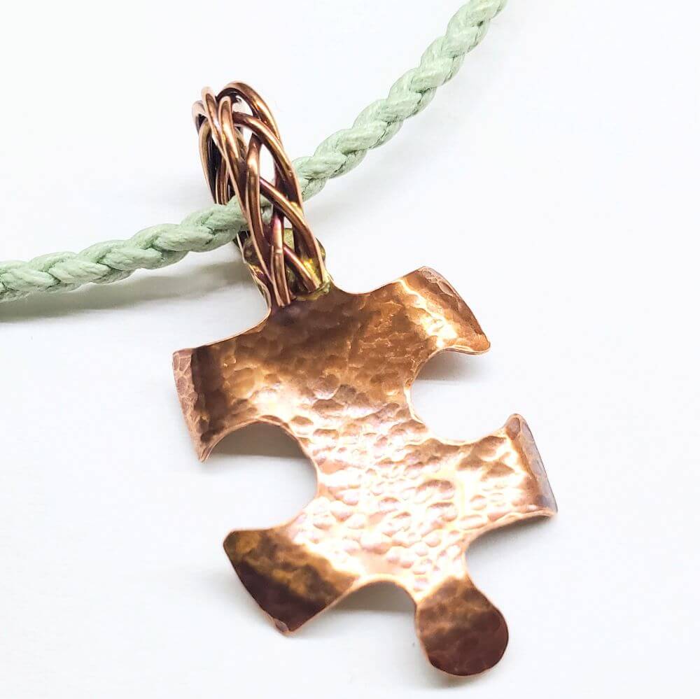 Copper puzzle piece pendant on a green braided cord against a white background