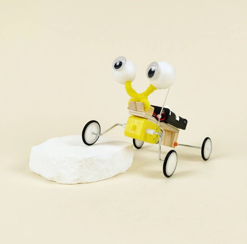 Toy robot figure with googly eyes and a small cart on a beige background