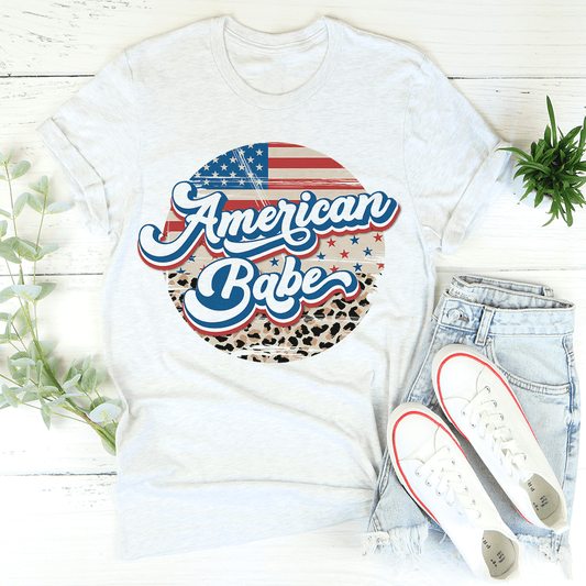White t-shirt with 'American Babe' design, light blue jeans, and white sneakers on a wooden surface.