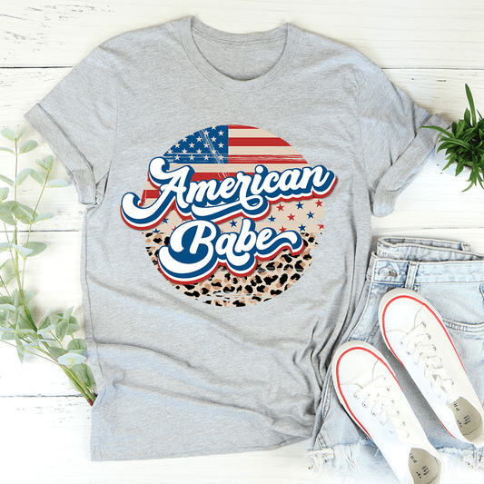 Gray t-shirt with 'American Babe' design on a white wooden surface with jeans and shoes.