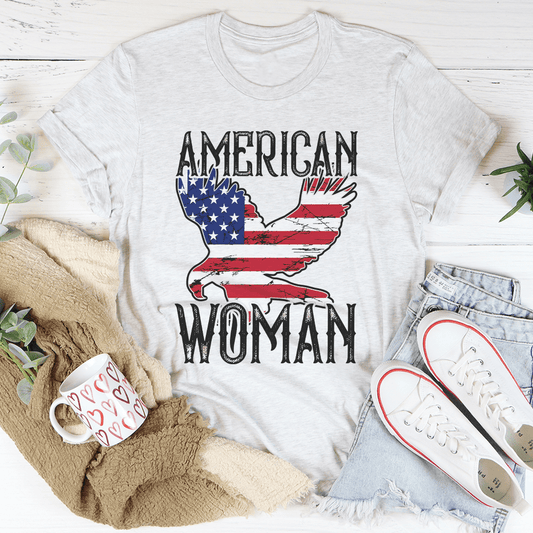 White t-shirt with 'American Woman' design featuring an eagle and American flag, placed on a white surface with casual items around.