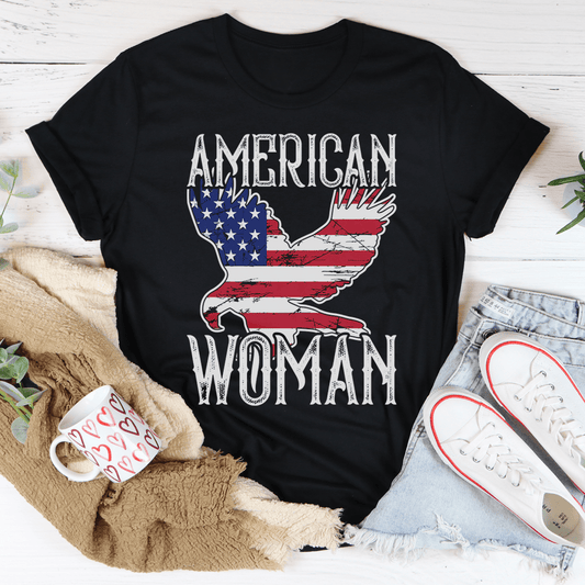 Black t-shirt with 'American Woman' design featuring an eagle and American flag, placed on a wooden surface with casual items.
