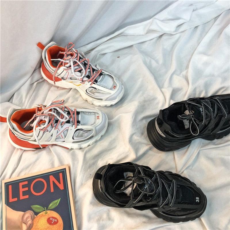 Two pairs of sneakers, one white with orange accents and the other black, on a light fabric background.