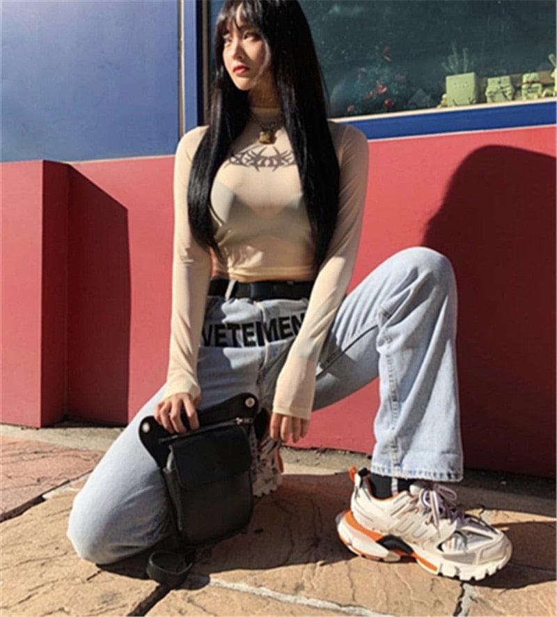 Person wearing a beige long-sleeve top and light blue jeans, sitting against a colorful wall.
