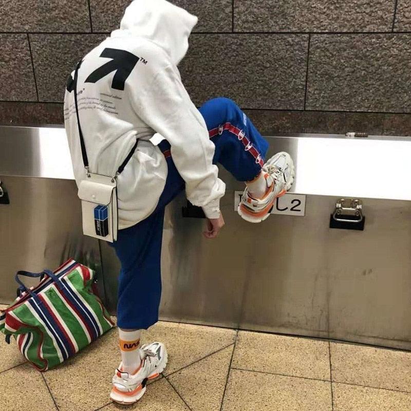 Person wearing a white hoodie and blue pants with colorful shoes, standing next to a striped bag on a tiled floor.