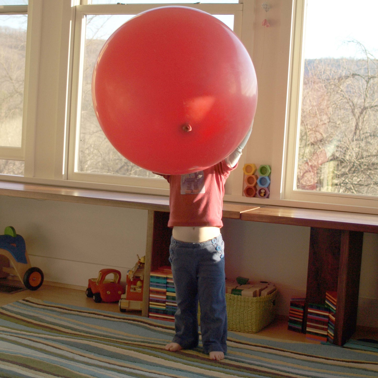 Child holding a large red balloon in a room with toys and a striped rug.