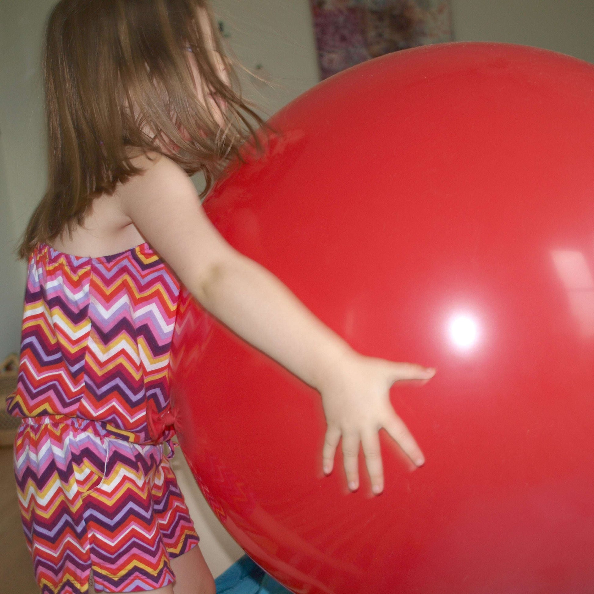 Child in a colorful dress holding a large red ball indoors.