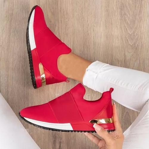 Red slip-on shoes with gold accents held by a hand on a wooden floor.