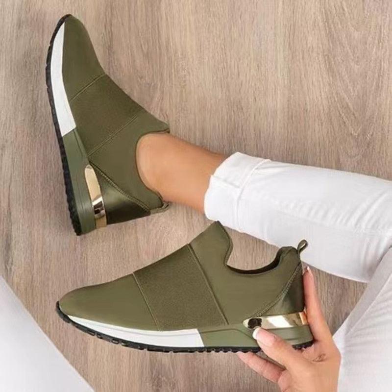 Green slip-on shoes with white soles worn by a person on a wooden floor.