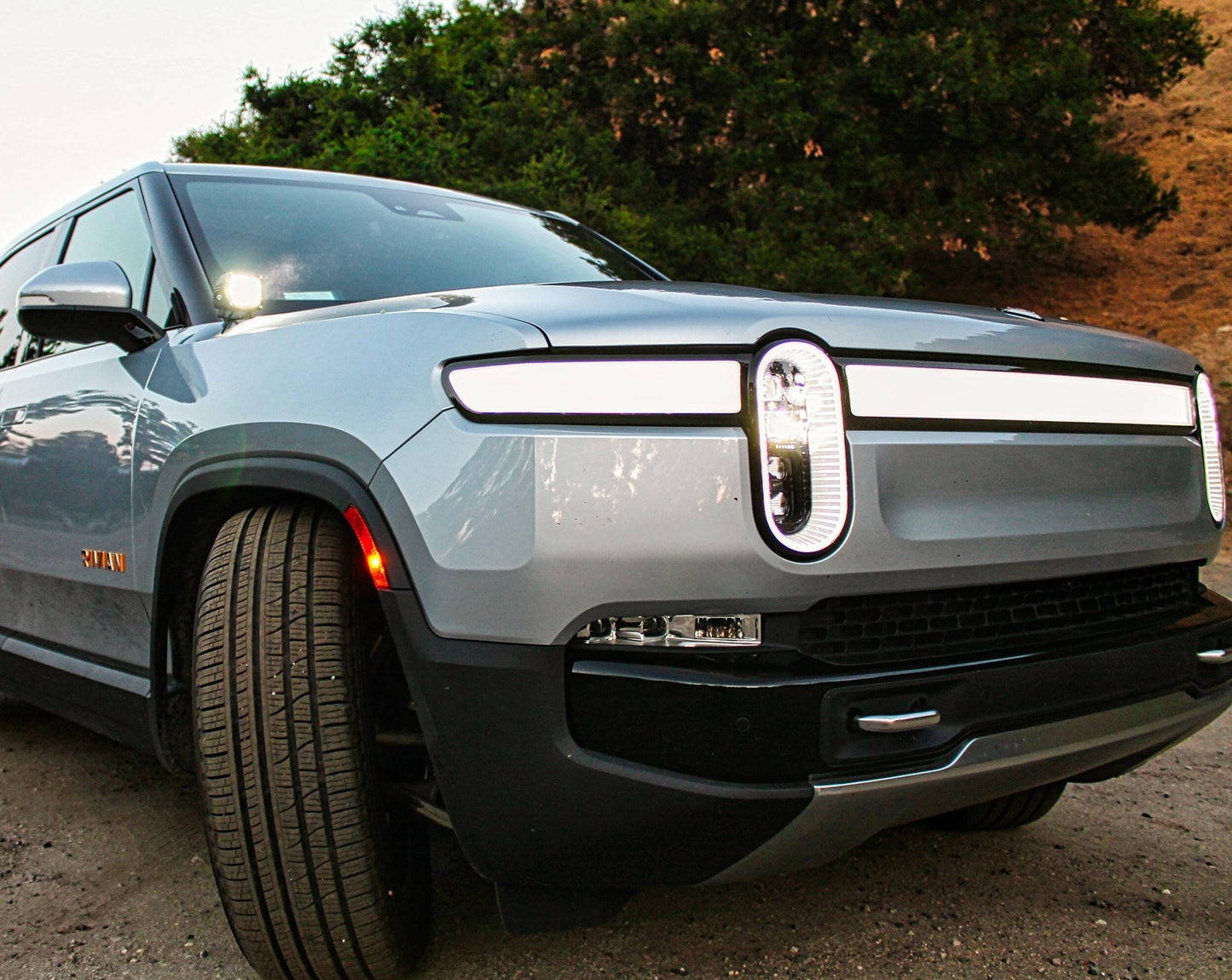 Front view of a silver Rivian electric vehicle on a dirt road with LED Light mounted on passenger side