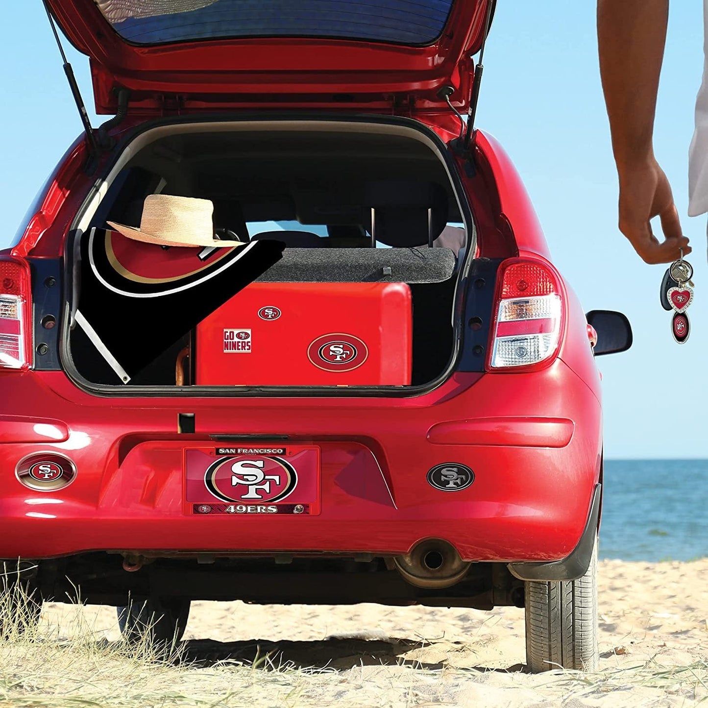 Red car with open trunk on a beach, containing a red cooler and a hat.