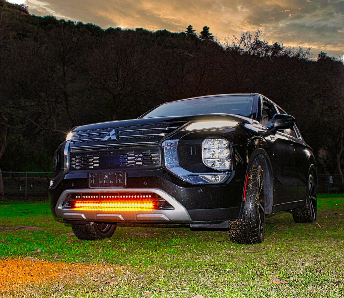 Black Mitsubishi car with illuminated amber front lights in a grassy area with trees in the background