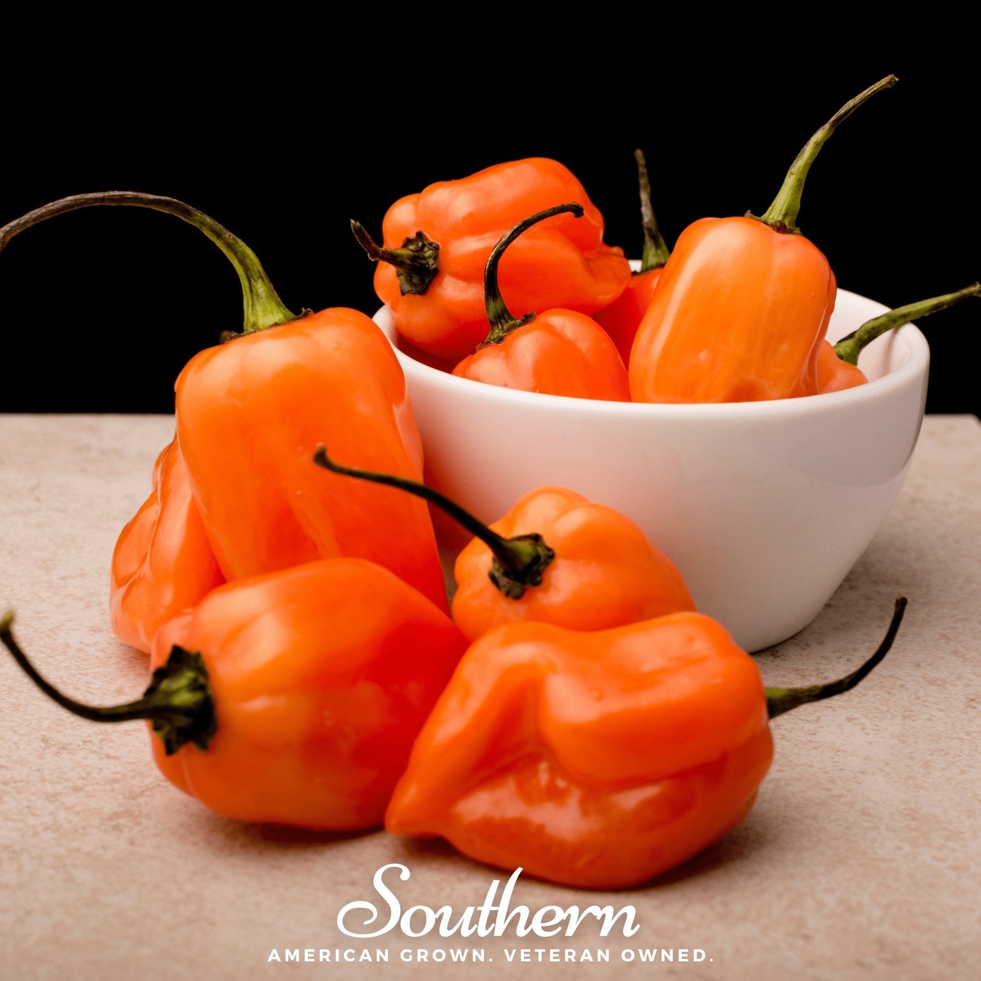 Orange peppers with green stems on a beige surface and a white bowl filled with peppers.