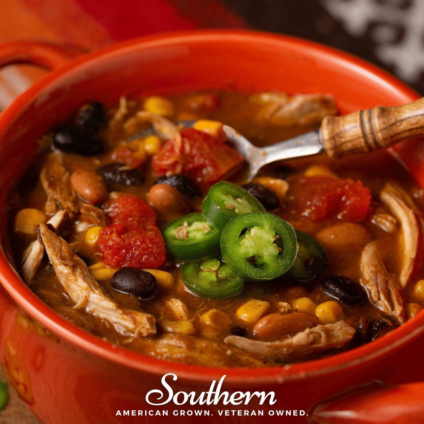 Red bowl of soup with jalapeños and visible branding on a wooden surface