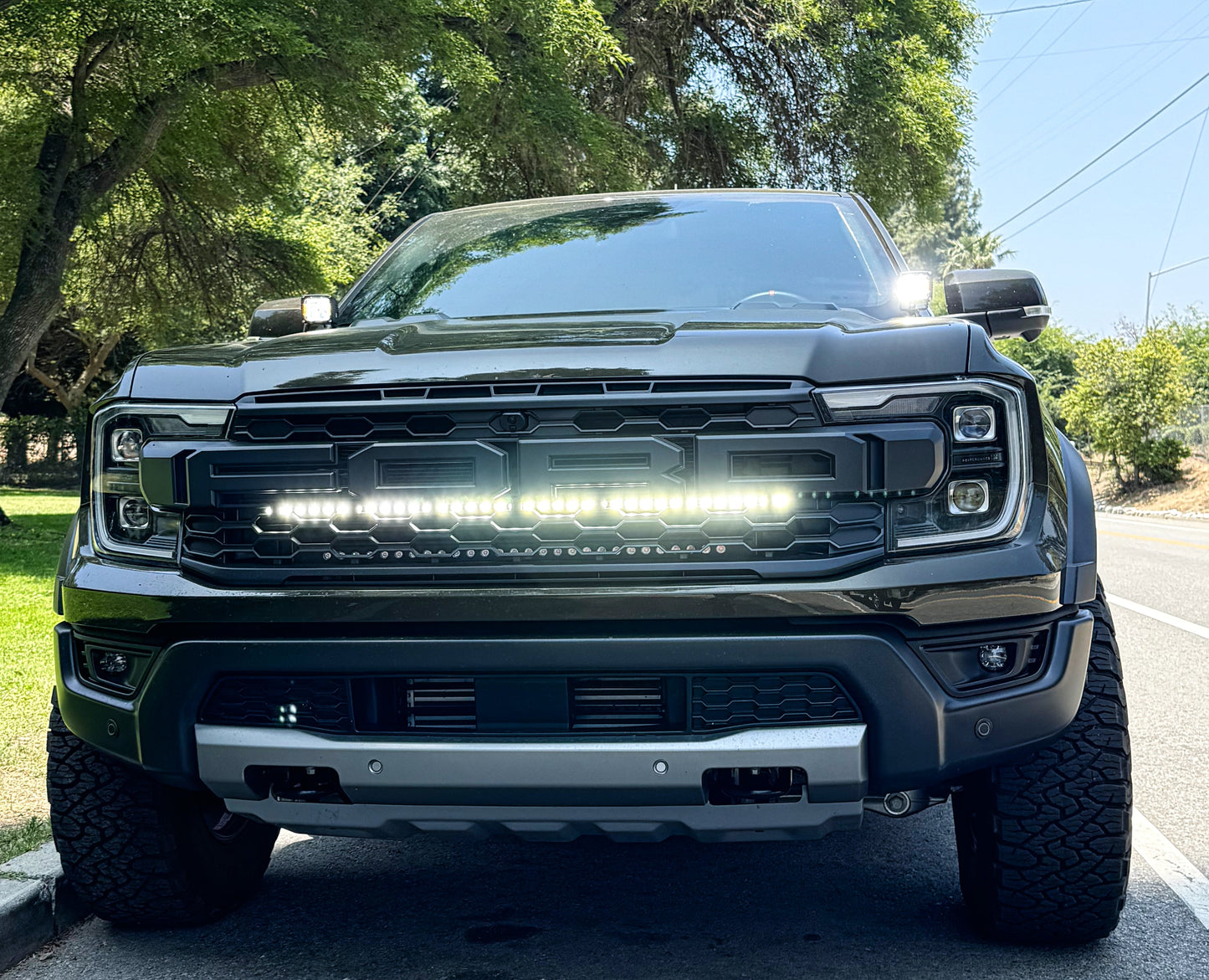 Front view of a black Ford F-150 Raptor truck on a road with trees in the background and LED Light bar mounted by the grille
