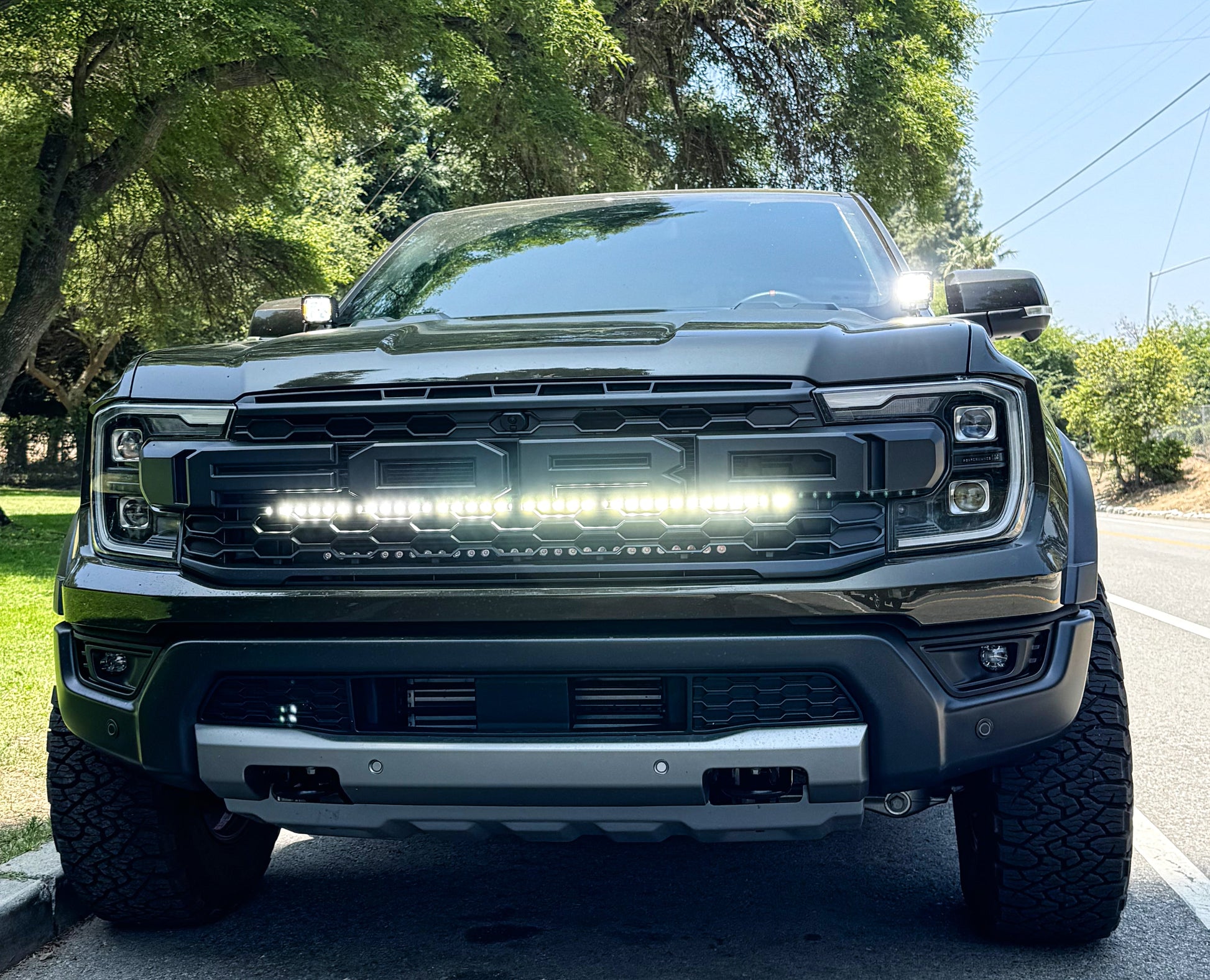 Front view of a black Ford F-150 Raptor truck on a road with trees in the background and LED Light bar mounted by the grille