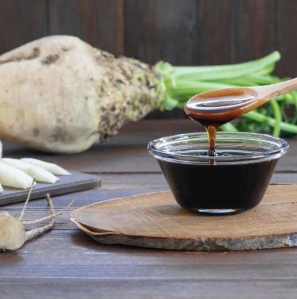 Wooden spoon drizzling dark liquid into a small container on a wooden surface with Sugar Beet roots and greenery in the background.