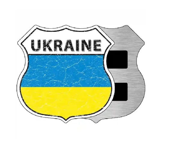 A shield-shaped Ukraine-themed emblem or patch with the word 'UKRAINE' at the top, a blue-and-yellow flag design in the center, and a brushed silver metal-like right side on a plain white background.
