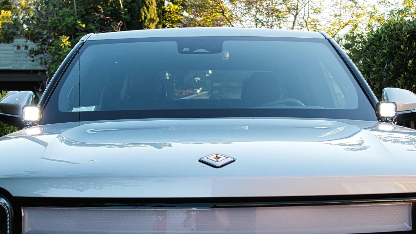 Front view of a white car with a visible logo, surrounded by greenery.